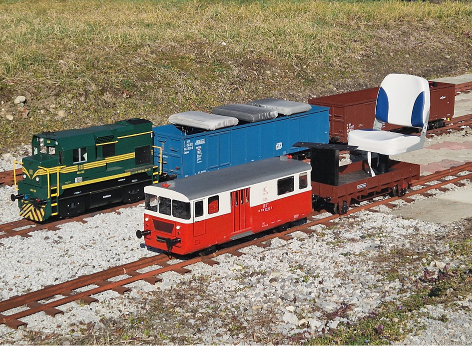 alt="Miniature red-and-white passenger train car, flatbed with seat, blue freight car, and green locomotive on narrow-gauge track outdoors"
